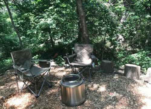 Two camping chairs beside a fire pit in a wooded area, surrounded by trees and wooden stumps.