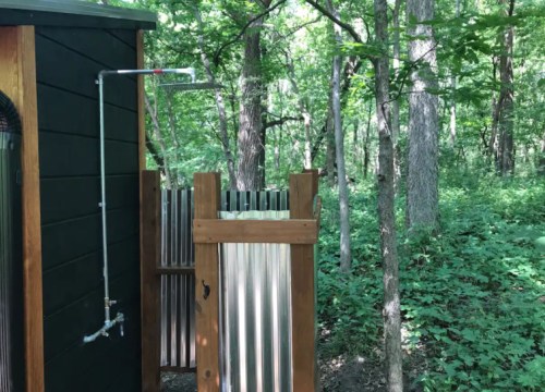 Outdoor shower surrounded by trees, featuring wooden and metal panels, in a lush green forest setting.
