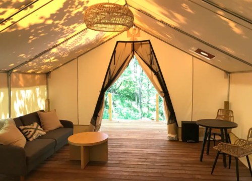 Cozy tent interior with a sofa, round table, and open entrance leading to a forest view. Warm lighting and wooden floor.