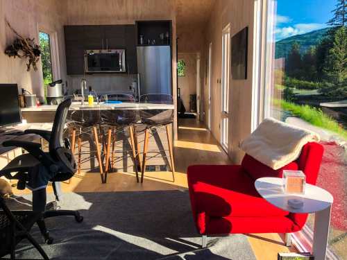 Bright interior of a modern kitchen and living area with a red chair, wooden table, and large windows showcasing nature.