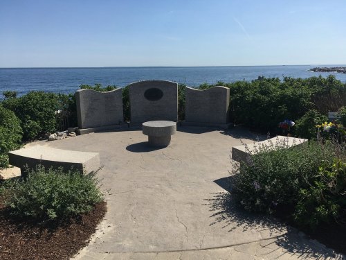 A serene memorial site by the ocean, featuring stone walls and a circular bench surrounded by greenery.