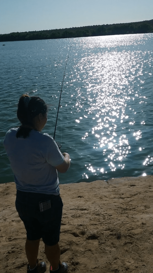 A person fishing by a shimmering lake on a sunny day, with trees in the background.
