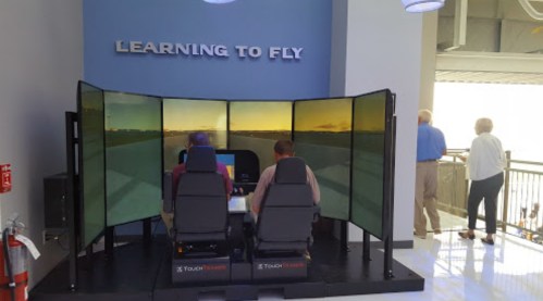 Two people sit in a flight simulator with large screens displaying a runway scene, while others observe nearby.