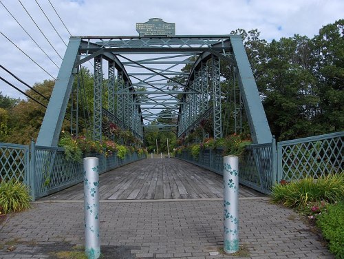 A green metal bridge with a wooden walkway, surrounded by trees and decorative flower planters, under a cloudy sky.