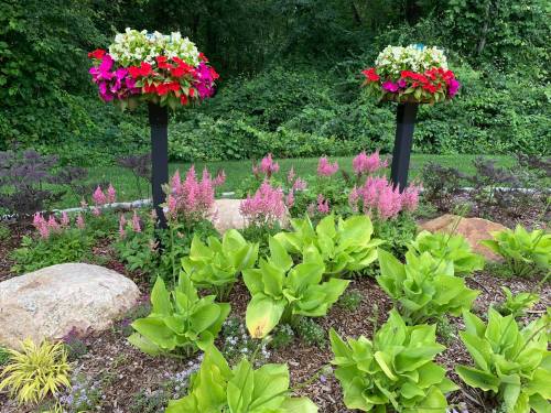 Two vibrant flower planters on posts surrounded by colorful flowers and lush green plants in a garden setting.