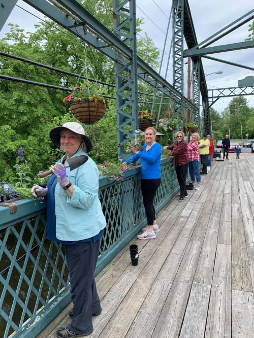 A group of people planting flowers on a bridge, surrounded by greenery, with hanging baskets and a cloudy sky above.