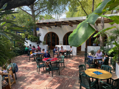 A vibrant outdoor café with green plants, colorful tables, and people dining in a sunny courtyard.