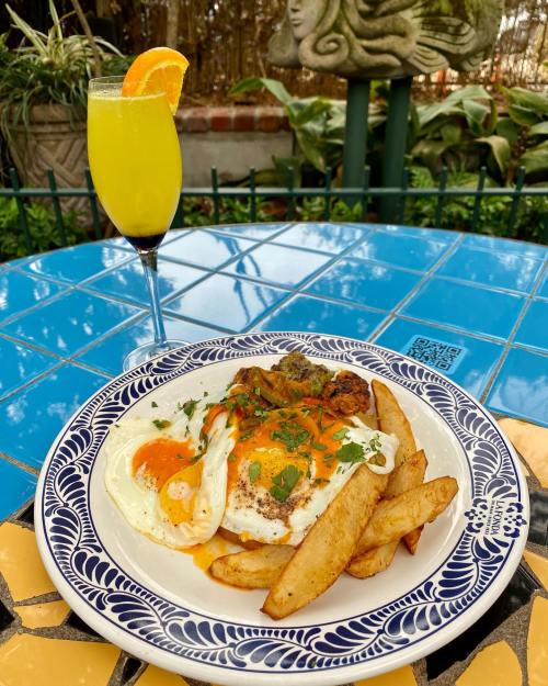 A plate of eggs with sauce and fries, accompanied by a yellow mimosa, on a colorful tiled table.
