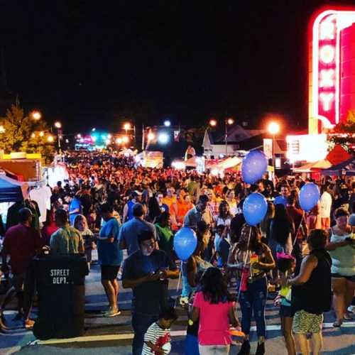 A bustling night street fair with crowds, food stalls, and colorful balloons under bright lights.