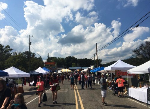 A bustling street fair with vendors under colorful tents, people walking, and a blue sky with fluffy clouds.