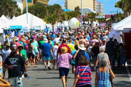 A crowded street fair with many people walking among tents and palm trees under a bright blue sky.