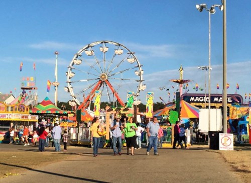 A lively fair scene with a Ferris wheel, colorful rides, and people walking along the midway under a clear blue sky.