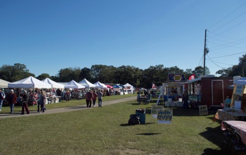 A bustling outdoor market with white tents, food trucks, and visitors enjoying a sunny day.