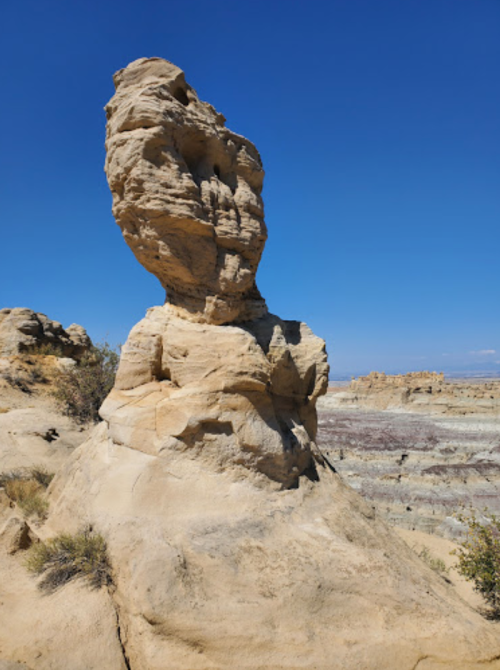 A tall, unique rock formation stands against a clear blue sky, surrounded by layered geological features.