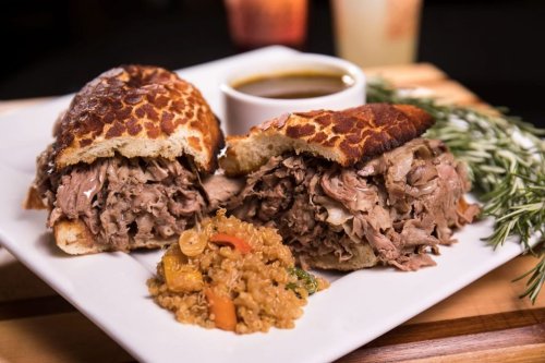 A plate featuring two hearty sandwiches filled with shredded meat, accompanied by a side of quinoa and a small bowl of sauce.