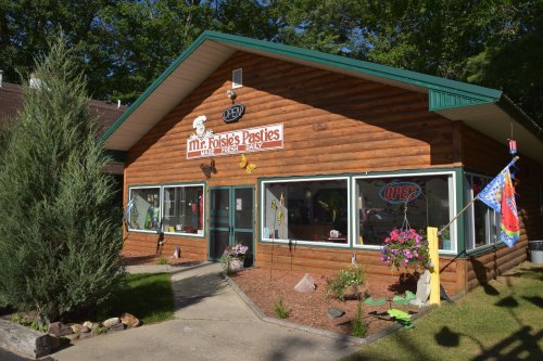 A rustic wooden building with a sign for "Mr. Josie's Pasties," featuring colorful flags and flower pots outside.