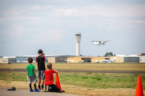 Three kids watch and photograph a plane landing at an airport, with a control tower visible in the background.