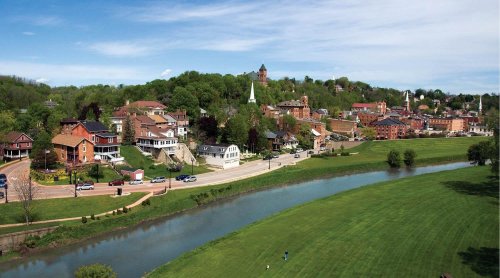 A scenic view of a small town along a river, featuring green hills, historic buildings, and a clear blue sky.