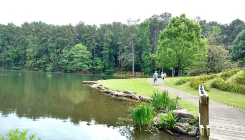 A serene lakeside scene with a walking path, lush greenery, and people enjoying the outdoors by the water.