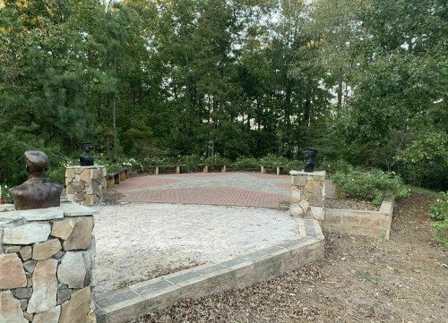 A stone patio surrounded by trees, featuring two bust sculptures and benches, with a garden area nearby.