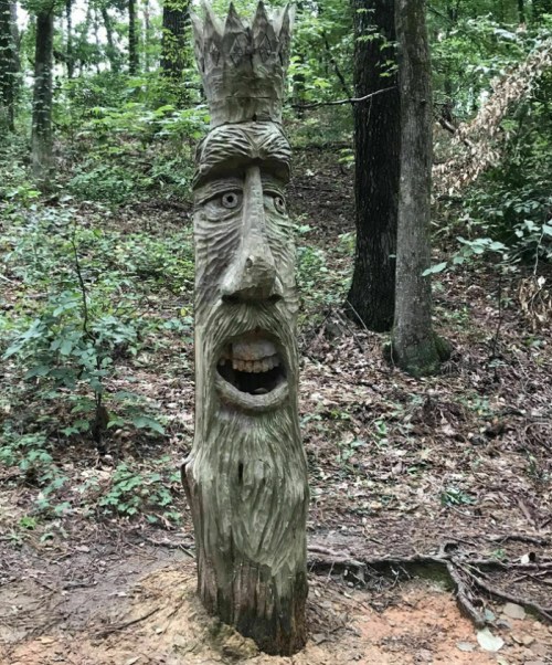 A carved wooden figure with a crown, featuring a surprised expression, stands in a forested area.