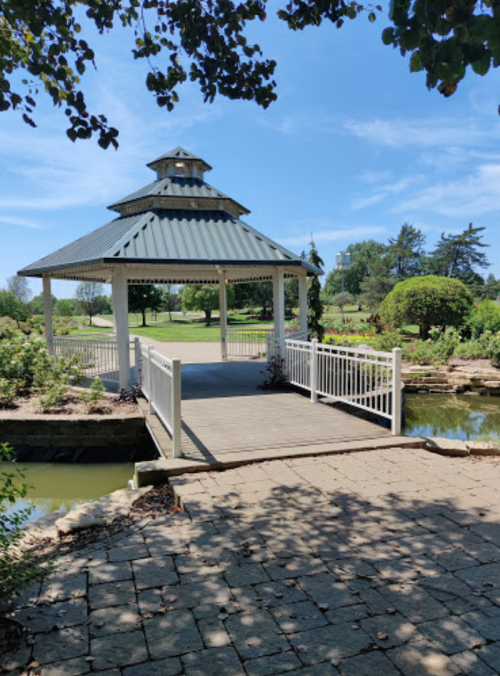 A white gazebo with a metal roof sits on a wooden deck, surrounded by greenery and a pond under a clear blue sky.