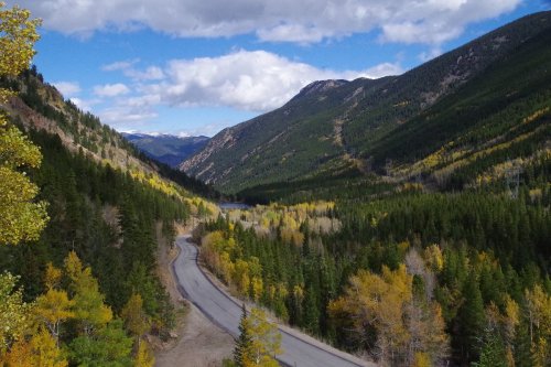 A winding road through a lush green valley surrounded by mountains and colorful autumn trees under a blue sky.