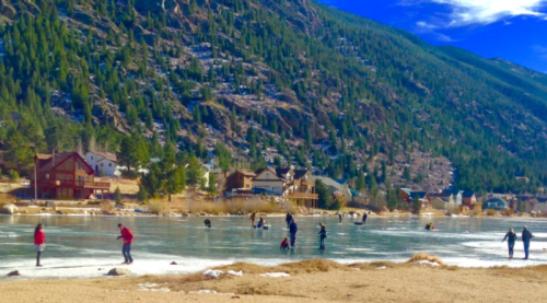 People ice skating and walking on a frozen lake, surrounded by mountains and houses in a scenic winter landscape.
