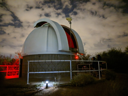 A small observatory dome under a night sky, with two people standing nearby, illuminated by red lights.