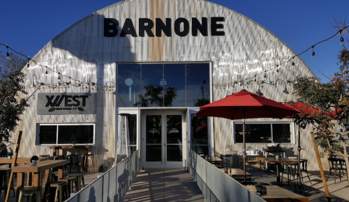 Exterior of a modern building labeled "BARNONE," featuring outdoor seating and red umbrellas under a clear blue sky.