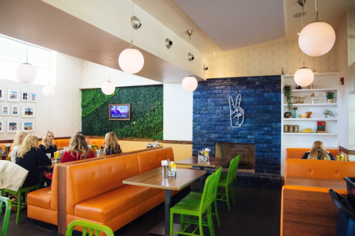 Bright restaurant interior with orange booths, green chairs, and a peace sign neon light on a blue brick wall.