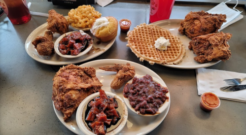 A table with two plates of fried chicken, waffles, macaroni and cheese, cornbread, and sides of beans and sauce.