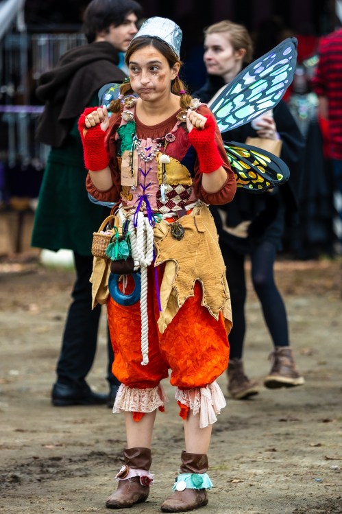 A young girl in a colorful, whimsical costume with butterfly wings, standing in a lively outdoor festival setting.