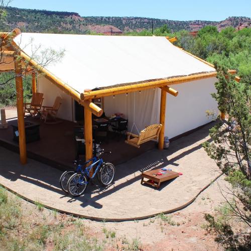 A cozy tent with a wooden porch, swing, and bike, surrounded by greenery and red rock formations.