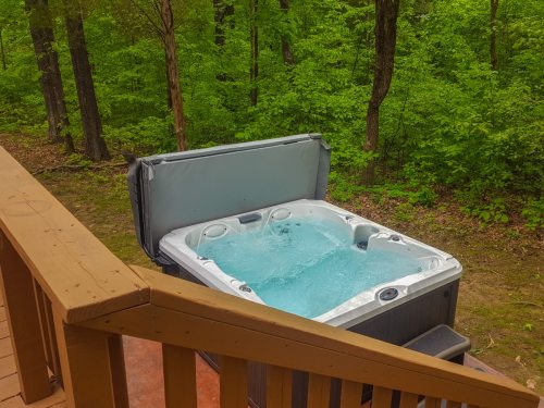 A hot tub with bubbling water sits on a wooden deck, surrounded by lush green trees.