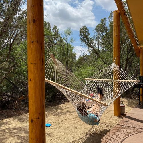 A person relaxes in a hammock surrounded by trees and a sandy area under a partly cloudy sky.