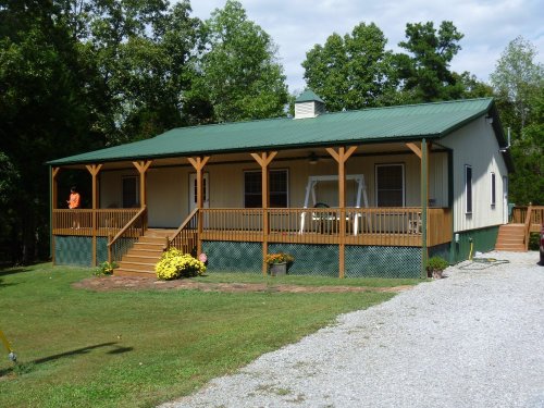A green-roofed house with a wooden porch, surrounded by trees and a grassy yard, featuring flower pots and a gravel driveway.