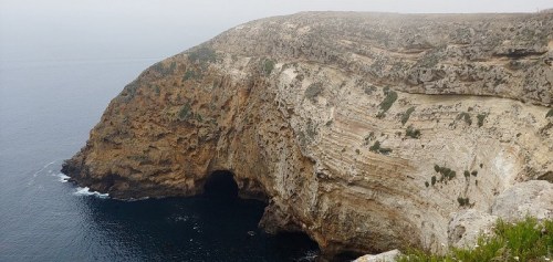 A rugged cliffside overlooking a dark cave and calm ocean waters, shrouded in mist.