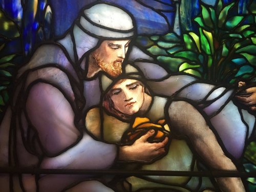 Stained glass depicting a man comforting a woman, surrounded by vibrant foliage and soft colors.