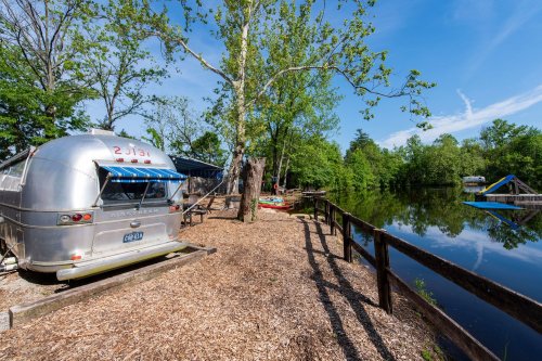 A silver Airstream trailer by a calm lake, surrounded by trees and a wooden fence, with kayaks visible on the water.