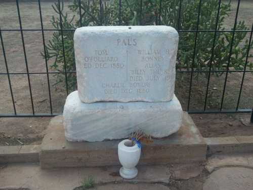 A weathered grave marker with inscriptions, surrounded by a fence and a small vase, set in a dirt area.