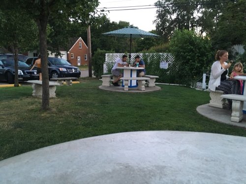 A park scene with families sitting at stone tables under an umbrella, surrounded by trees and grass.