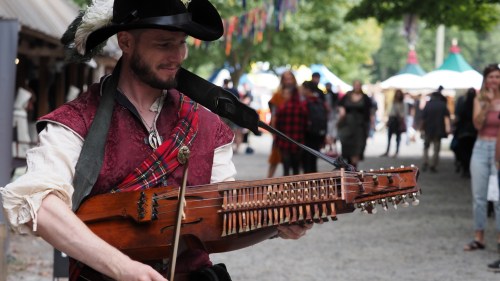 A man in a hat plays a stringed instrument at a lively outdoor festival, with people and colorful decorations in the background.