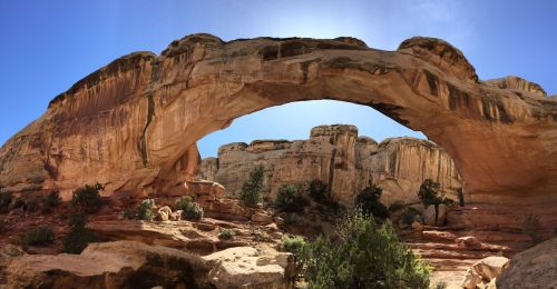 A natural stone arch rises above a rocky landscape under a clear blue sky, surrounded by rugged cliffs and vegetation.