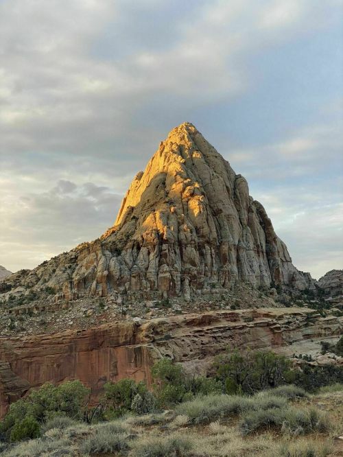 A towering rock formation bathed in golden light, surrounded by rugged terrain and a cloudy sky.