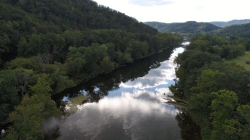 A serene river flows through lush green hills under a cloudy sky, reflecting the landscape in its calm waters.