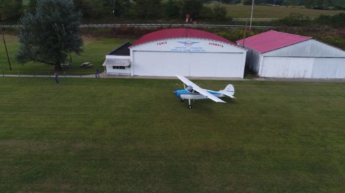 A small airplane parked on a grassy field near a hangar and a few people in the background.