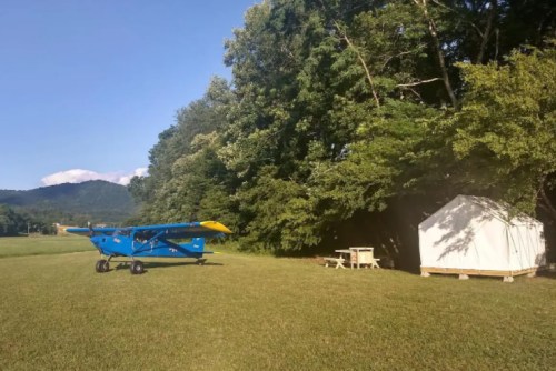A blue airplane parked on a grassy field next to a white tent and picnic table, surrounded by trees and mountains.