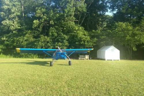 A blue aircraft with a yellow tip parked on a grassy area near a white shed and surrounded by trees.