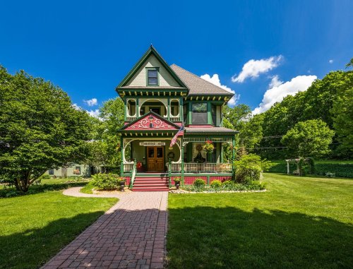 A charming Victorian-style house with a green exterior, red accents, and a well-kept lawn under a blue sky.